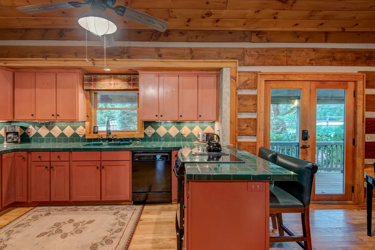 This kitchen-focused image highlights green tiled countertops, wooden cabinets, and bar stools, with a view that suggests access to the outdoors. The space is well-equipped and designed for both functionality and warmth, featuring ample counter space and cozy touches like a patterned rug.