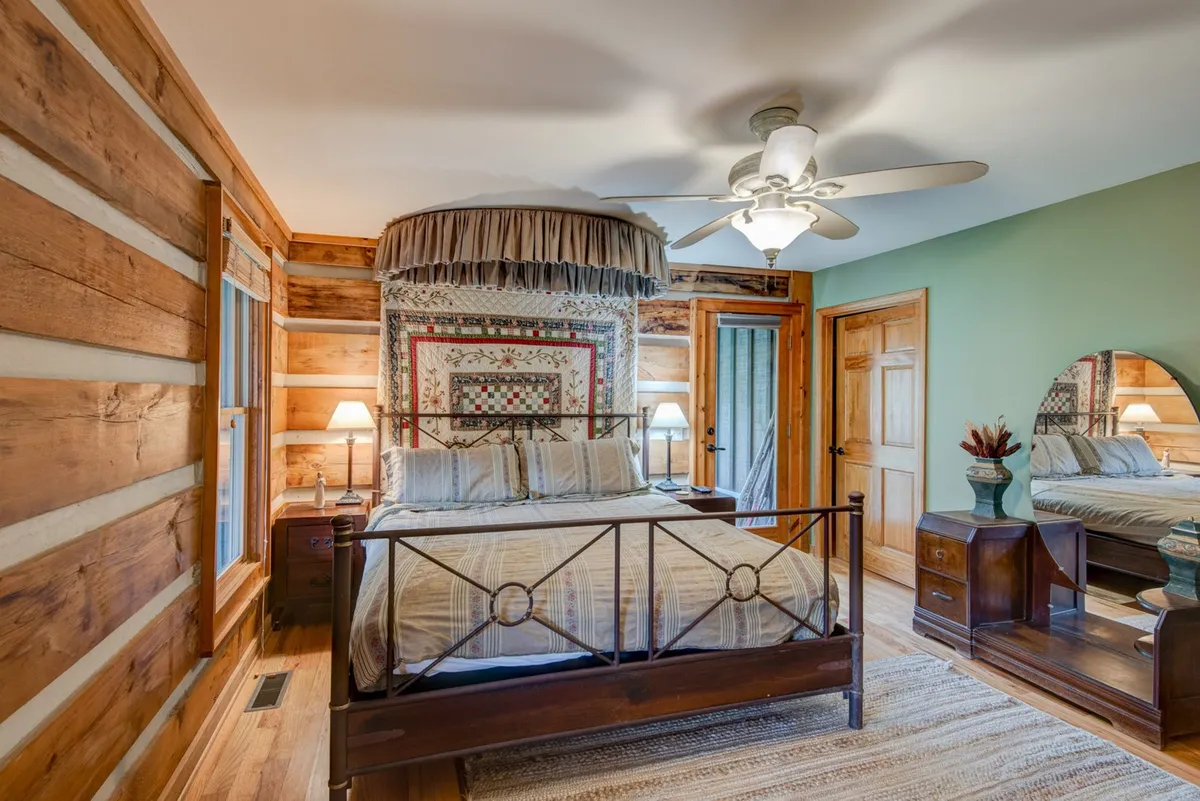A cozy bedroom featuring a comfortable bed with a quilted wall hanging above, warm wooden walls, and a mirrored dresser. The room exudes a homey atmosphere, inviting guests to relax, read, or unwind after a day of adventure.