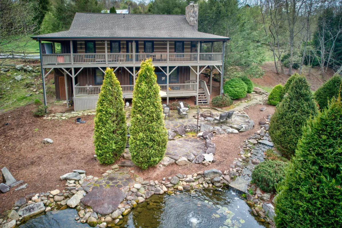 The image showcases the serene exterior of a single-family home in Boone, North Carolina, nestled among two tall conifer trees. A stone pathway leads to a tranquil pond that reflects the golden hues of sunset. A cozy patio area with inviting chairs overlooks the water, perfect for relaxing or gathering with loved ones. The lush lawn glistens with morning dew, and a fire pit hints at magical starry nights outdoors.