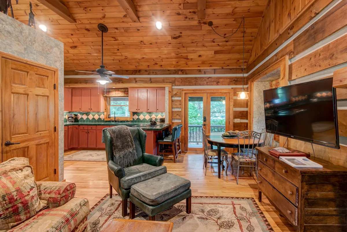 This photo captures an open-plan living area where a comfortable seating area transitions seamlessly into a kitchen and dining space. Visible are the wooden beams overhead, colorful kitchen cabinetry, and a dining table with a view of the outdoors. The overall ambiance is warm and homely, providing a sense of connected living spaces.