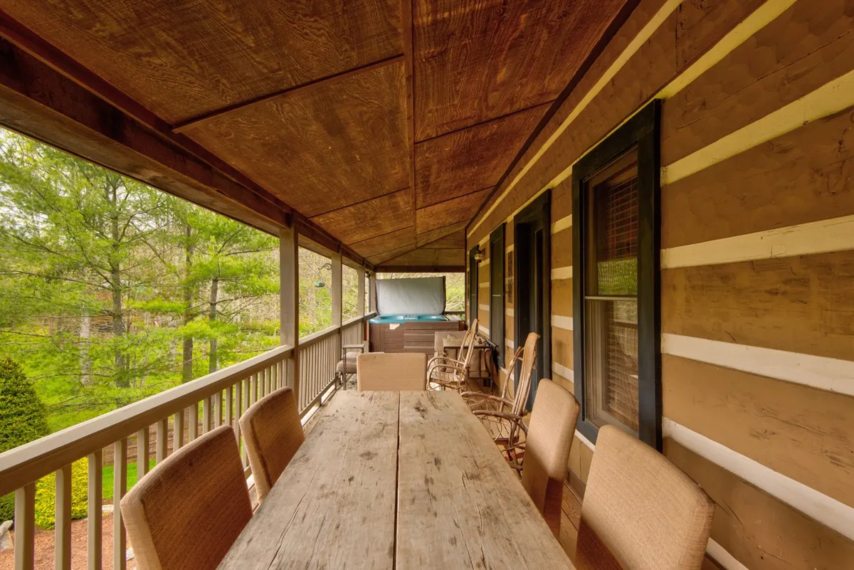 A wooden outdoor dining table and chairs set under overhead beams, surrounded by greenery. The scene feels casual and welcoming, inviting guests (and their pets) to enjoy meals and relaxation outside.