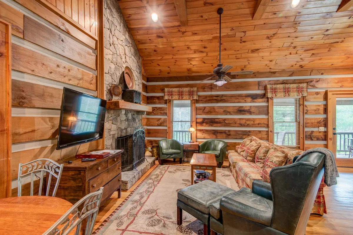 This image showcases a spacious great room with a prominent stone fireplace as a central feature. Large wooden beams accent the ceiling, and plush sofas form a comfortable seating area perfect for gatherings or relaxing. A decorative rug and tasteful decor create a warm, inviting atmosphere. The television is visible, hinting at entertainment options.